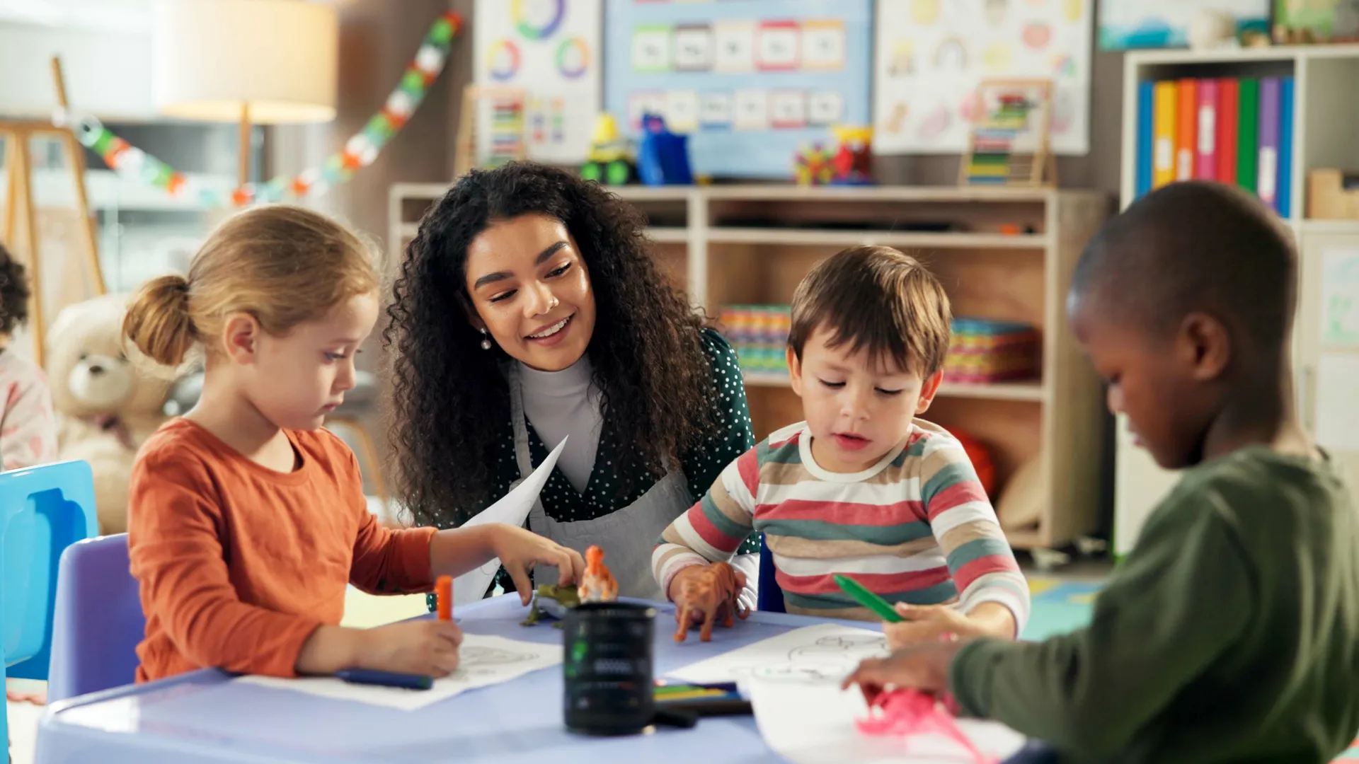 A woman with long curly hair kneels next to a table with three young, diverse students in a classroom setting. 