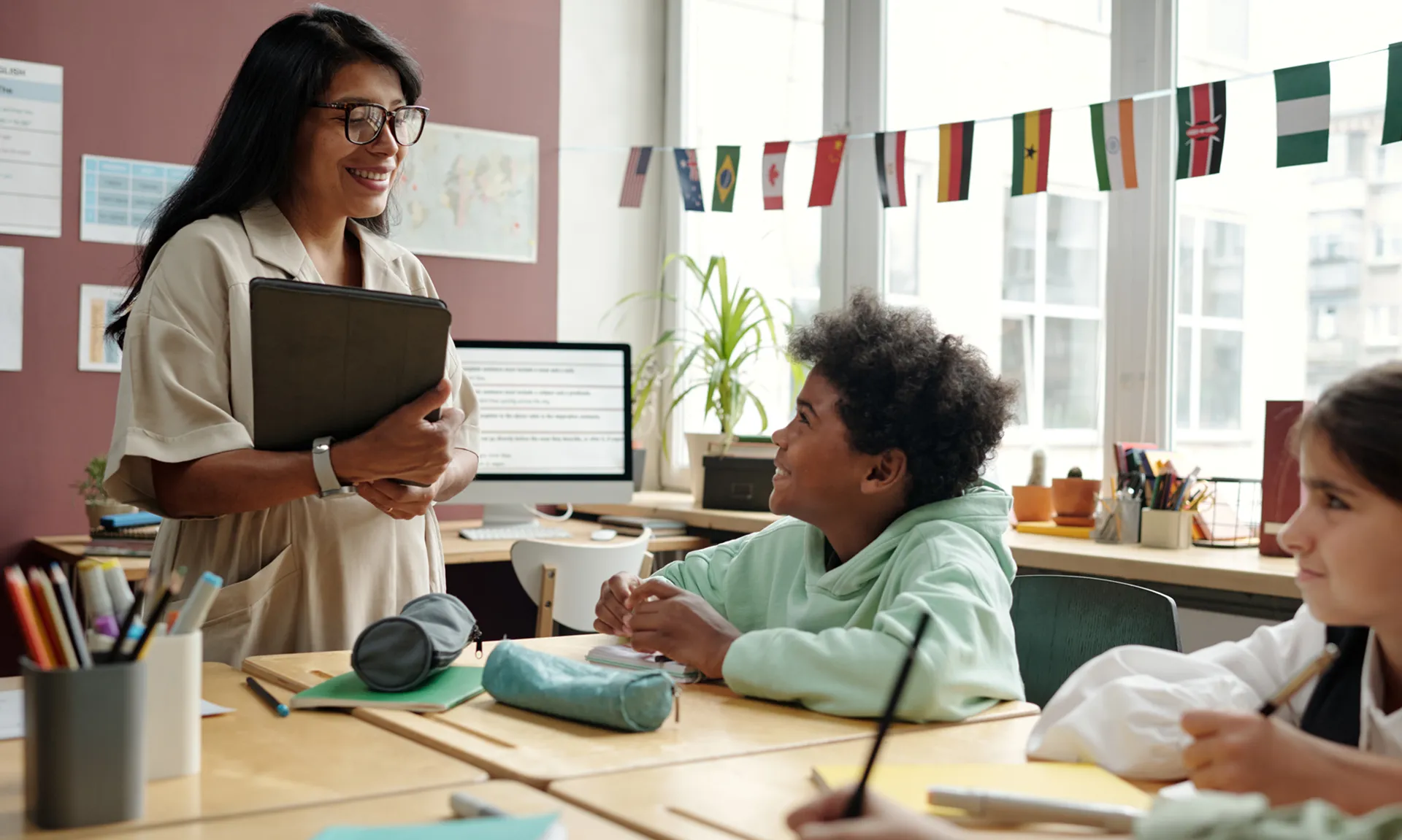 A woman wearing glasses and a white top holds a tablet in her arm while smiling at two students who are sitting at a desl. 