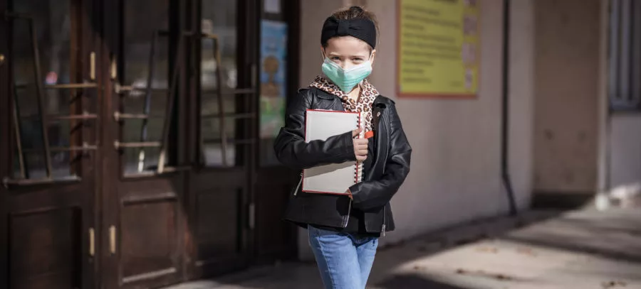 Scared young child with protective mask and books in her hands exiting school