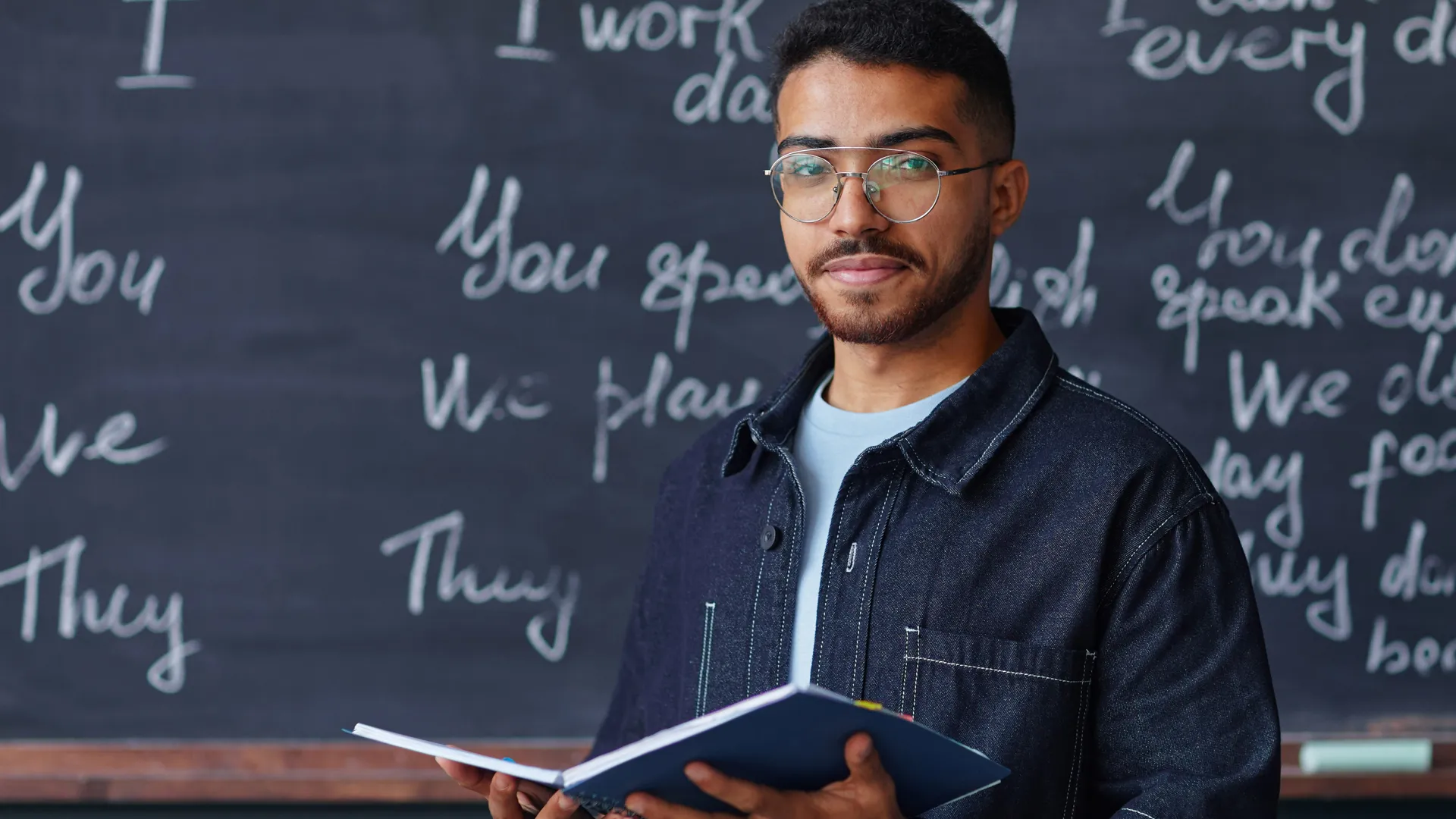 A man wearing glasses holds an open book in his hands and stands in front of a chalkboard with writing on it.