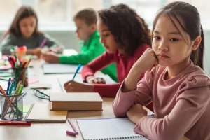 A student in a classroom posed with her fist to cheek looks off into the distance while other students work in the background. 