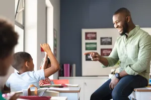 A male teacher sits on a desk while pointing to a student who's raising his hand.