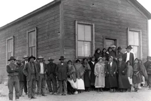 A black and white 1920s photo showing a large group of people in front of a log building.