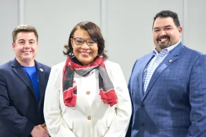 A woman in a white blazer and black and red scarf wrapped around her neck stands between two men in suit jackets.