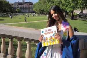 A woman with long brown hair smiles softly at the camera while holding a book in front of her that reads "All Are Welcome."