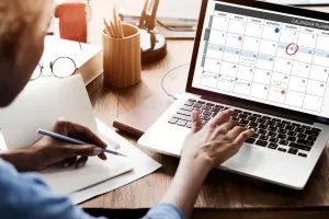 A woman sits behind a desk, holding a pen in one hand and working at a desk on a laptop displays a calendar.
