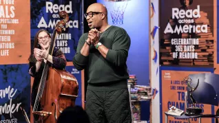 Author Kwame Alexander stands with hands clasped and addresses an audience of students with bassist Amy Shook in the background.