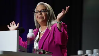NEA Executive Director in a pink blazer addresses RA delegates from a lectern
