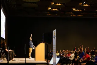 A male NEA staff member stands at a lectern with his hand raised asking the audience a question during a professional delegate training