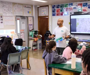 A male teacher stands in front of a classroom, addressing a group of students seated at their desks.
