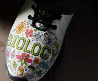 A person wearing a shoe with the word "biology" spelled across top and surrounded in colorful science cells.