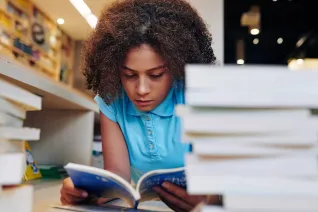 teen girl reading a book in the school library next to a stack of books on the desk