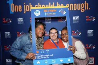 Three women of color standing in front of a backdrop that says, "One Job Should Be Enough," and holding up a dark blue social media frame labeled "NEA ESP Bill of Rights" in orange letters