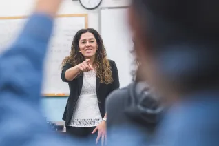Female teacher with white top, black blazer, and long brown hair points to call on a student in a classroom