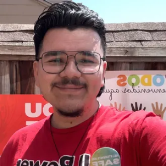 A man wearing glasses and a red shirt stands in front of a sign, smiling and looking towards the camera.