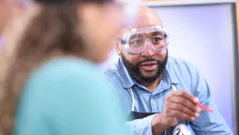 An emerging teacher wearing lab goggles supervises students conducting a science experiment