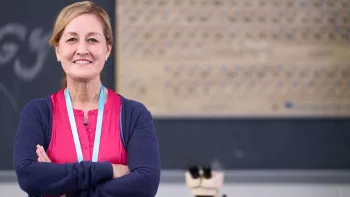 A middle-aged female teacher stands in front of a chalkboard with her arms folded