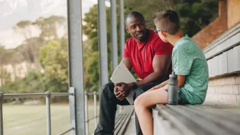 Black male coach in a red shirt sits on bleachers outside counseling a white male students in a green shirt