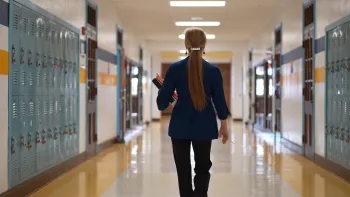 Female teacher with long ponytail walking down empty school hallway away from the camera. Hallway is lined with lockers