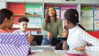 Black female teacher stands next to a table of students in the classroom during a discussion