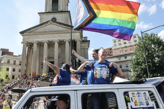 People cheerfully waving a rainbow flag while standing on top of a vehicle during a festive event.