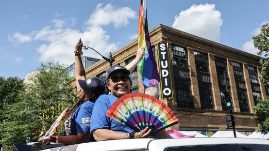 Two women in a car wave colorful rainbow flags, expressing joy and support for the LGBTQ+ community.