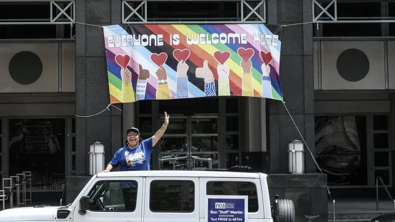 A woman waving from the top of a white truck, standing in front of an Everyone is Welcome Here sign.