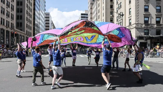 A street scene with several people gathered, holding a large, colorful parachute banner.