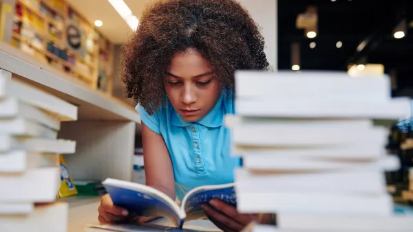 teen girl reading a book in the school library next to a stack of books on the desk