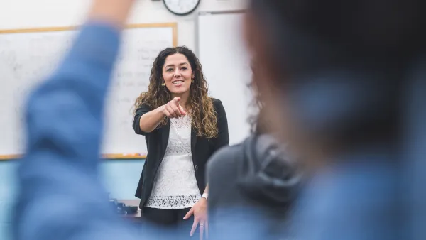 Female teacher with white top, black blazer, and long brown hair points to call on a student in a classroom