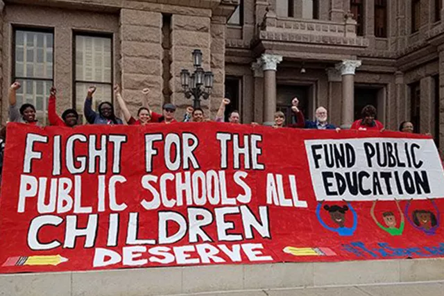 People with fists in the air hold a large painted banner saying "fight for the public schools all children deserve."