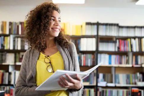 Woman with a yellow shirt and grey cardigan with curly brown hair smiles in a library while holding a book.