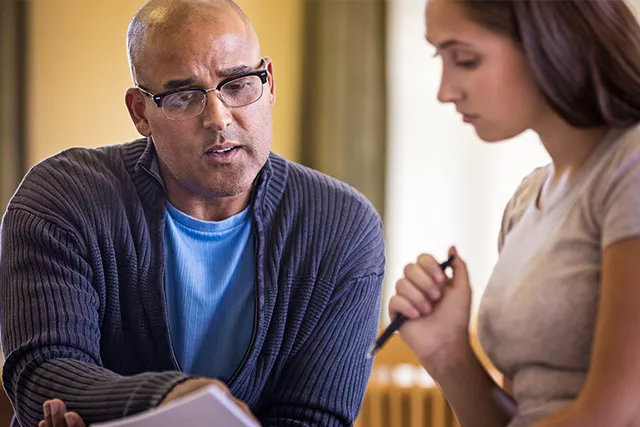 Bald man in glasses points to a document held by a woman with long brown hair with a pen.