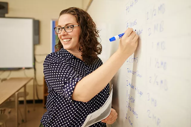 Teacher with brown curly hair and glasses smiles while writing on a white board.