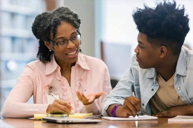 A teacher wearing glasses and wearing a pink long sleeved shirt sits with a student with a jean jacket and t-shirt.