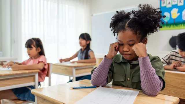 A young black girl sits in a classroom with other children and is starting at a paper on her desk, appearing frustrated