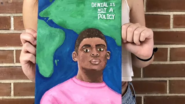 Student holding a painting of a man in front of a the earth that says "Denial is Not a Policy"