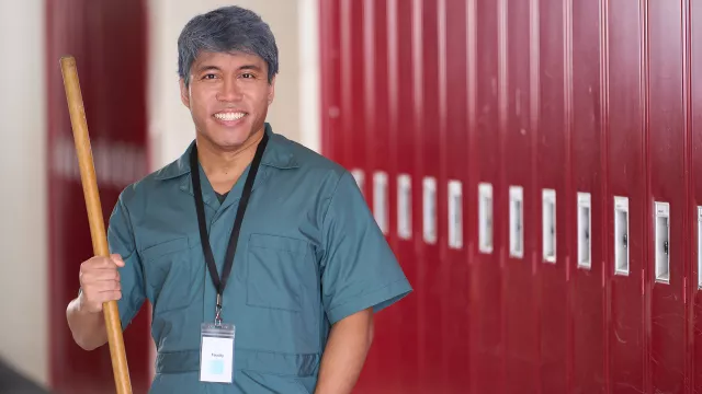 male school custodian in uniform stands in front of a wall of red lockers