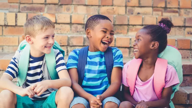 3 young children sit outside school wearing backpacks and smiling
