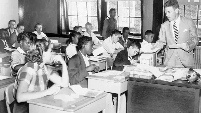 A white male teacher hands out papers in a 1960s classroom of black and white high school students