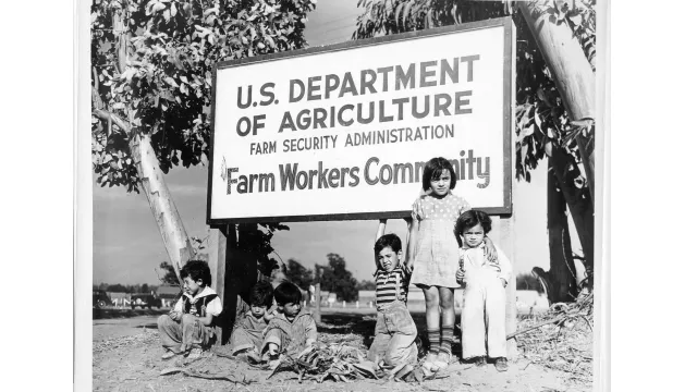 Group of children posing under sign that reads "U.S. Department of Agriculture Farm Security Administration Farm Workers Community" circa 1941