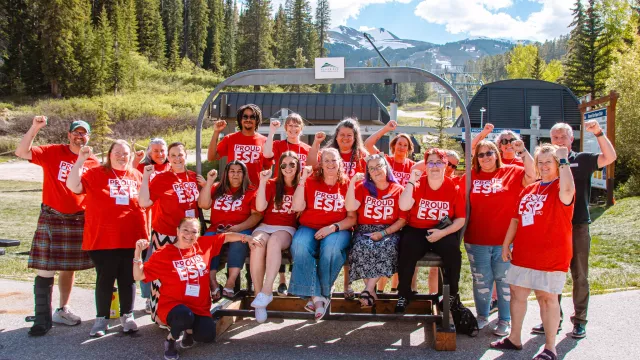 Educators in red shirts saying "ESPs" sit in front of a ski lift with their fists in the air.