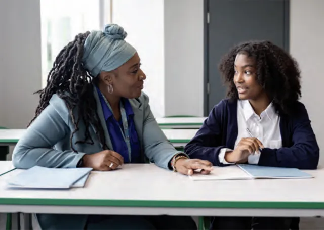 Black female teacher and student sit at a desk and discuss schoolwork