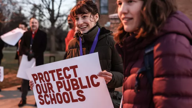 Woman stands in front of school with sign that says "protect public schools."