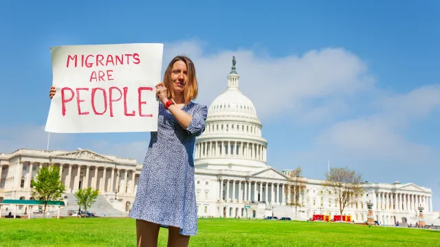 A person standing in front of the U.S. Capitol holding a sign that says, "Migrants are People."
