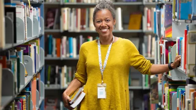 African American librarian in yellow shirt stands in the library stacks and smiles at the camera