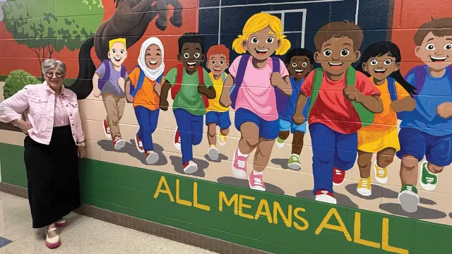 Retiree Joni Richman Watson in front of a student mural with words that say All Means All