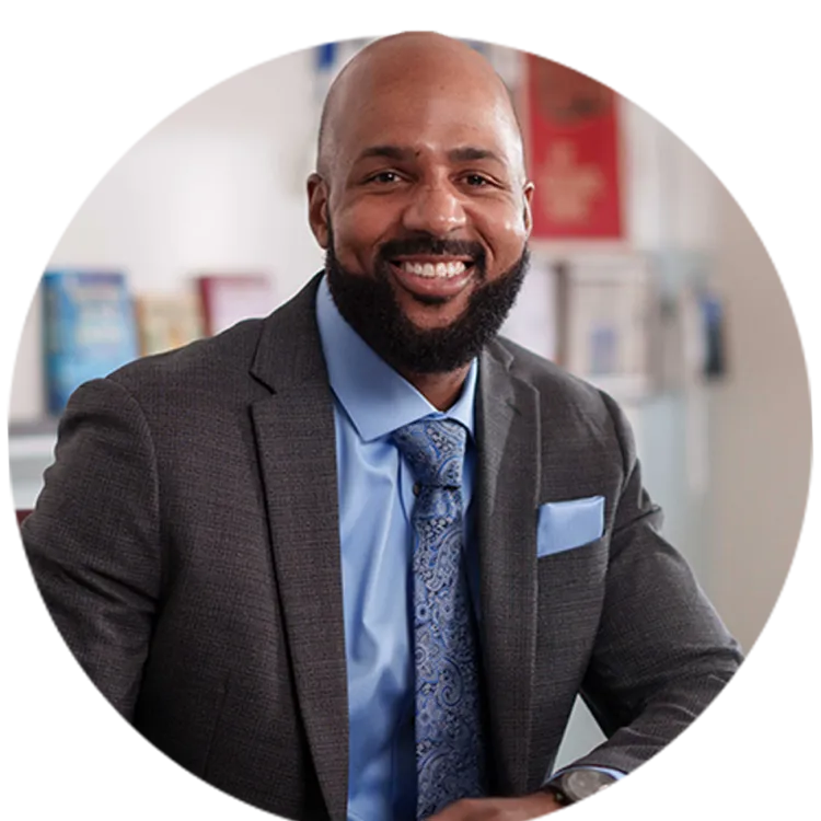 Leon Smith wearing a suit and tie smiles for the camera while seated at his desk in his classroom