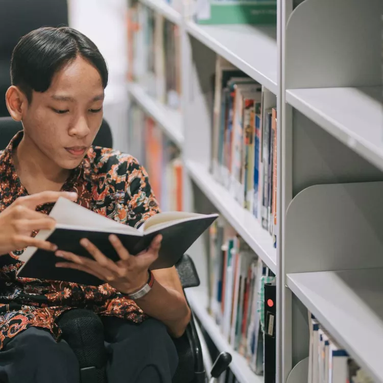 A teenage boy in a wheelchair browses books in a school library