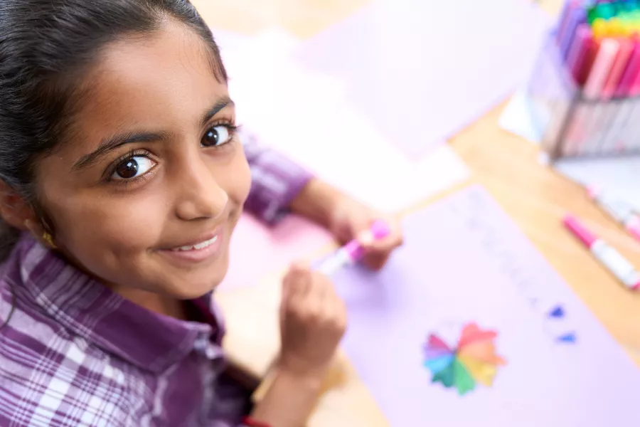 young girl seated at table looks up from her drawing and into the camera